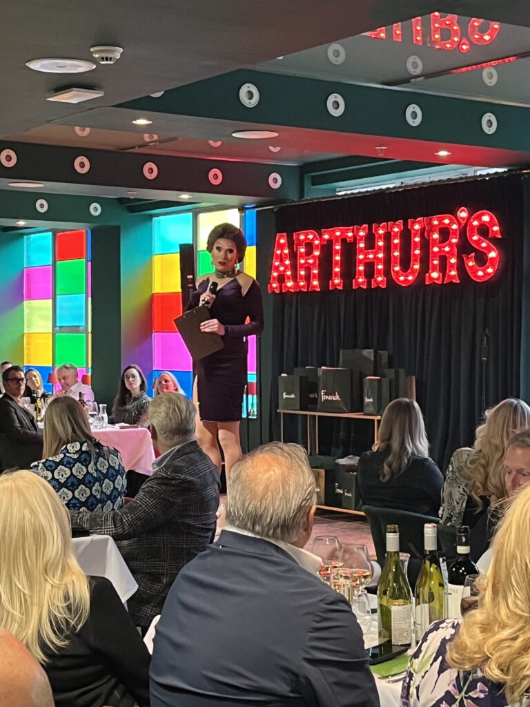 A compere standing on a stage with a sign behind them which says 'Arthur's' and a crowd of people sitting watching.