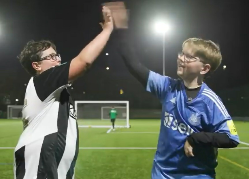 Two white boys aged about 13, one blonde and one with brown hair, both wearing glasses, high five on a football pitch. One is wearing a Newcastle United home shirt and the other us wearing an away shirt