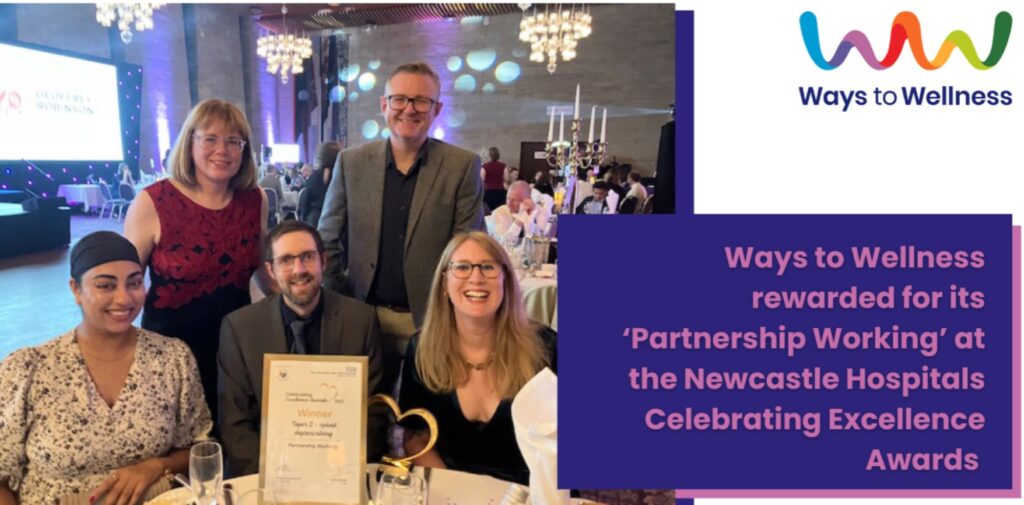 A diverse group of professionals, three sat at a white tableclothed table with a framed certificate and gold hear-shaped trophy in front of them, and two stood behind are all smiling at the camera.