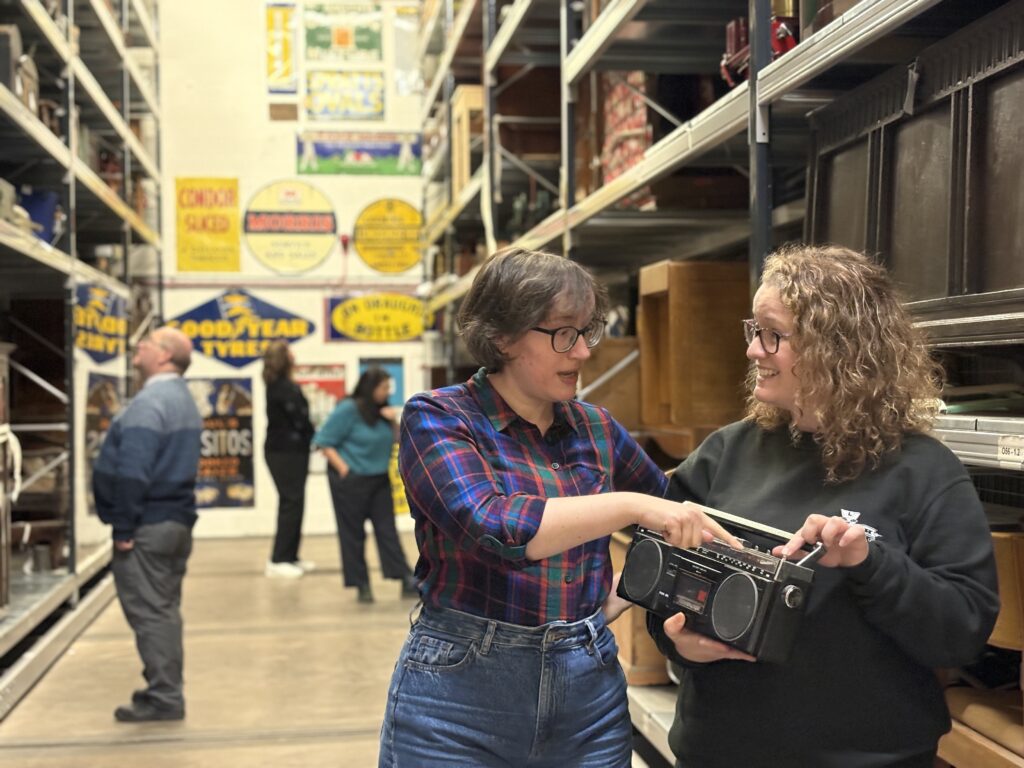 Two people stand in the open store at Beamish Museum. One is holding a radio, the other is pointing at it. There are people in the background looking at the objects on shelves.