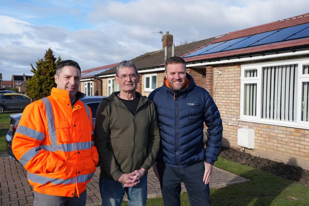 Photo of Jonathan Horner, from REGEN Group, believe housing tenant Peter Tickle, and David Taylor, from believe housing, outside homes near Durham which have recently had home energy efficiency upgrades, including new roofs with solar panels which can be seen in the background.